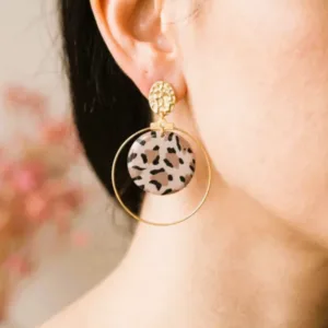 Une femme portant une boucle d'oreille pendante composée d'une puce d'oreille dorée texturée et d'un grand anneau doré entourant un disque plat au motif léopard imprimé.
