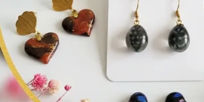 Collection de boucles d'oreilles artisanales en résine présentées sur un support blanc, incluant des modèles en forme de cœur et des pendentifs.