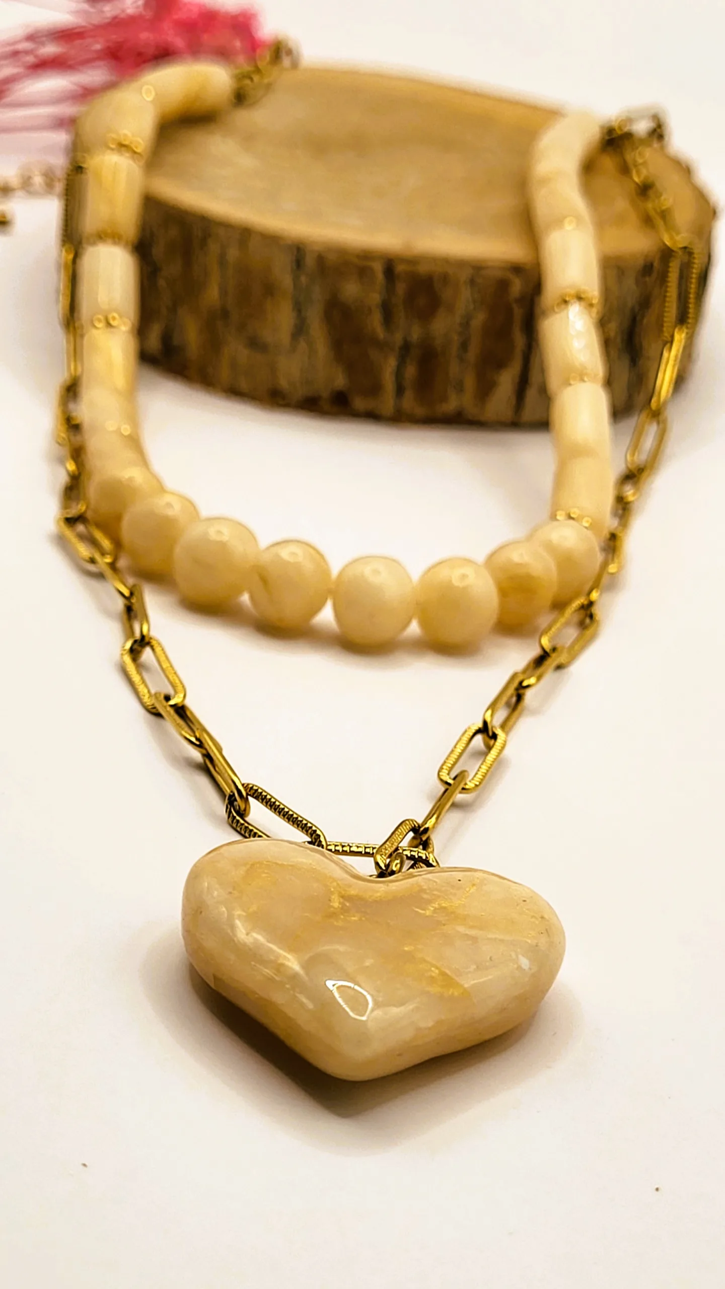 Collier double rang composé d'un brin de perles nacrées oblongues et d'une chaîne dorée en maille rectangle avec un grand pendentif cœur marbré beige et or, posé sur une tranche de bois.