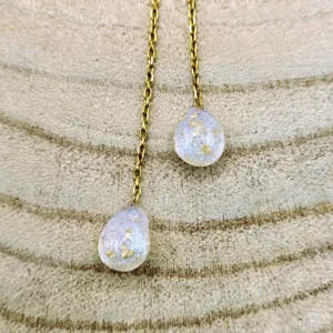 Gros plan sur les deux pendentifs en forme de gouttes en résine, ornés de délicates inclusions dorées, suspendus à une chaîne dorée.